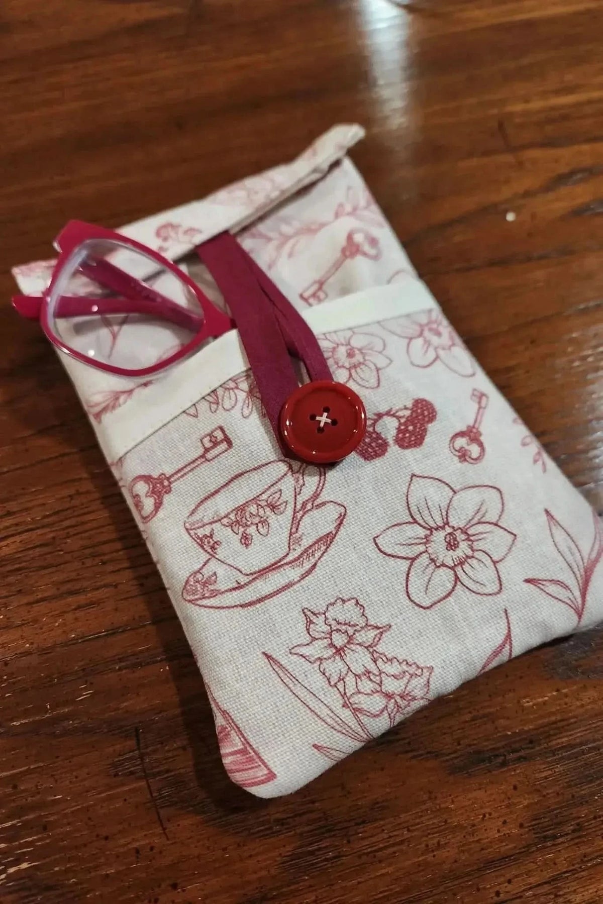 Red fabric pouch with tea cups, keys, and flowers print, holding pink glasses.