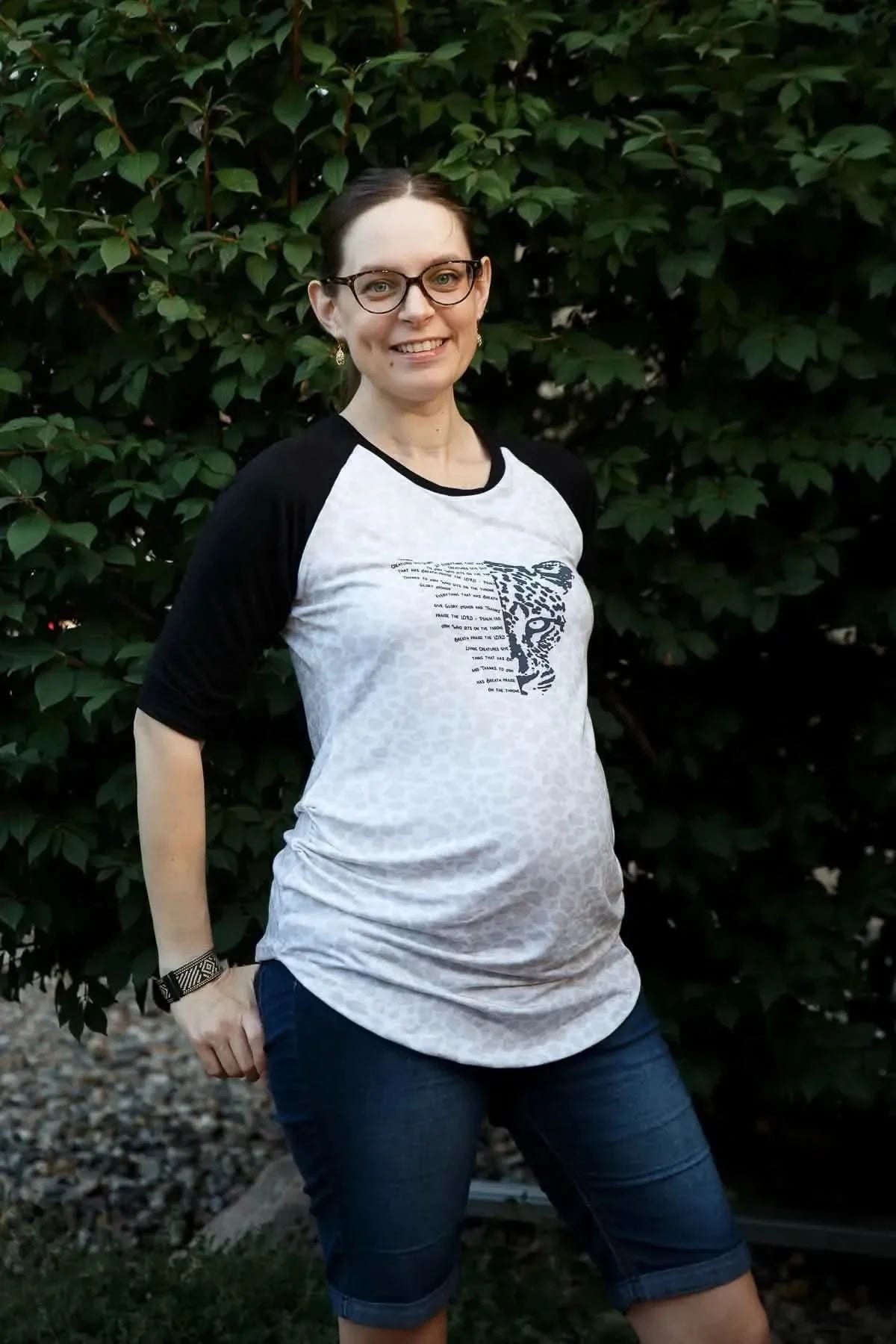 Pregnant woman wearing a white raglan T-shirt with black sleeves and Creatures Give Glory panel print.