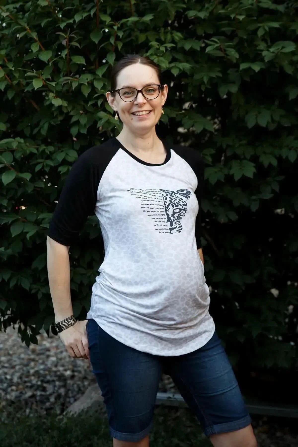 Pregnant woman wearing a white raglan-sleeve shirt with black creature graphic and text.