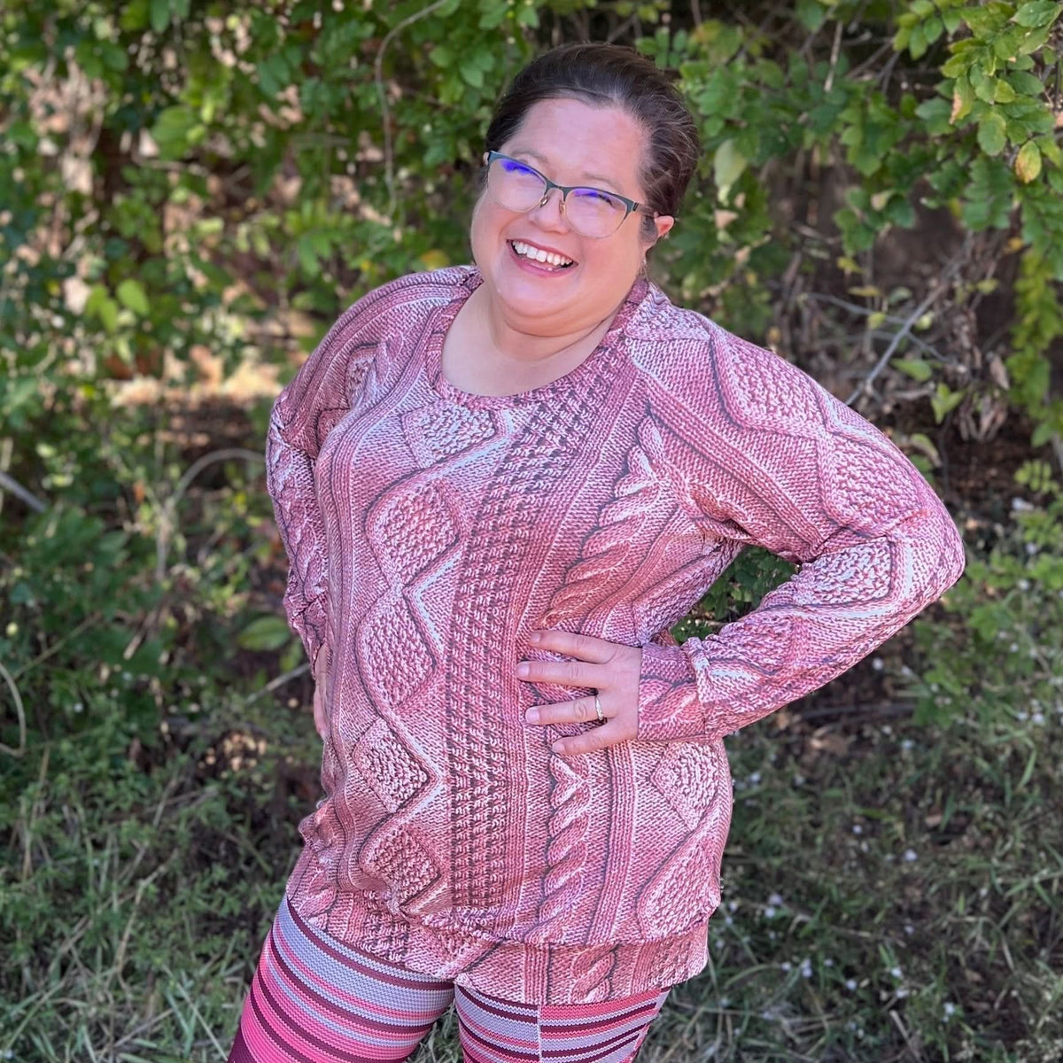 A woman smiles, wearing a rose cable knit top and striped leggings.