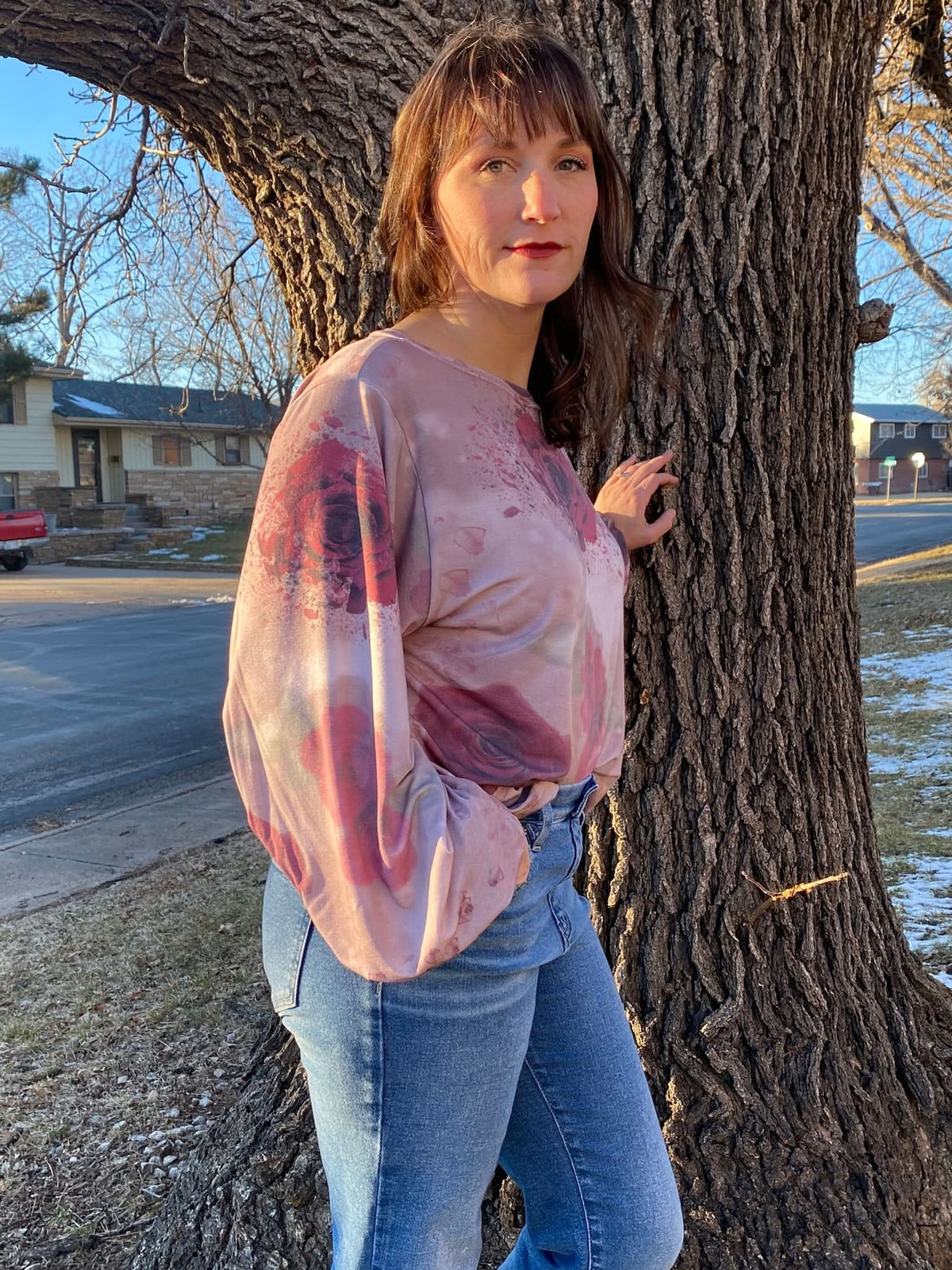 A woman wears a pink top with a shattered rose pattern, leaning against a tree.