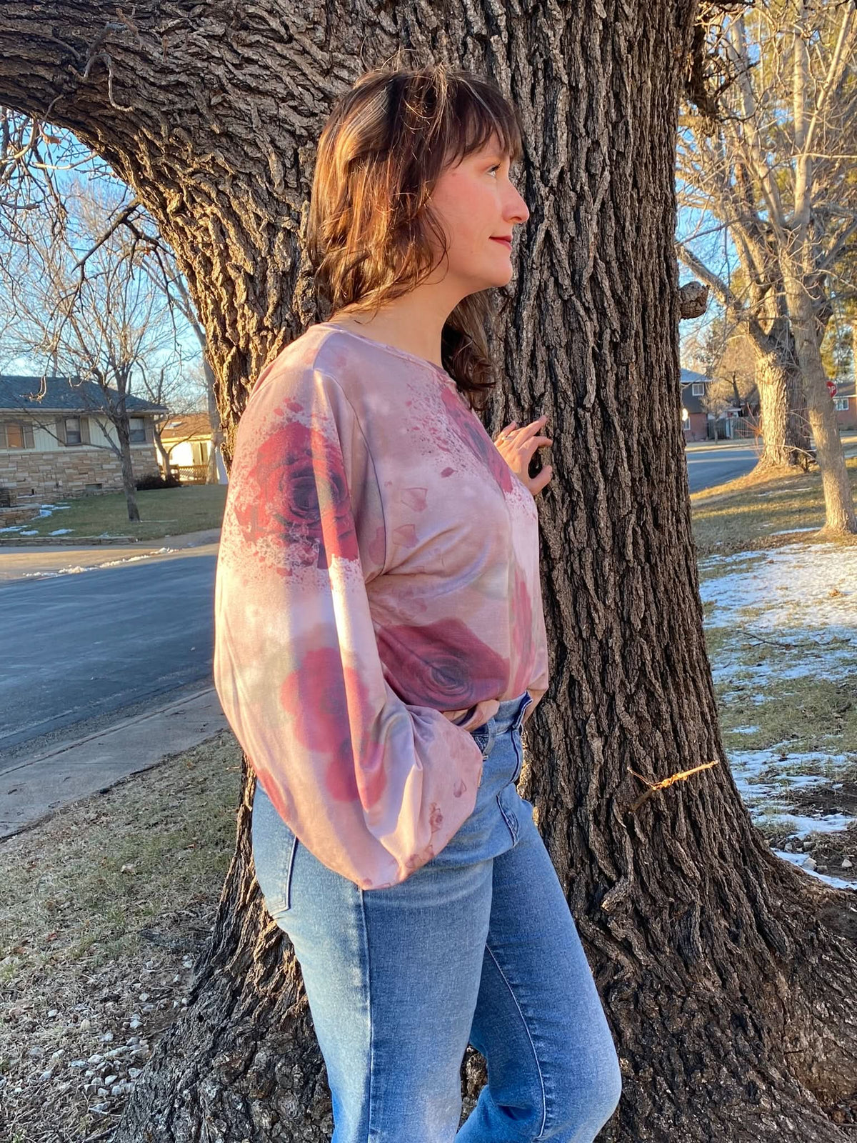 A woman in a pink rose-patterned top and jeans leans against a tree.