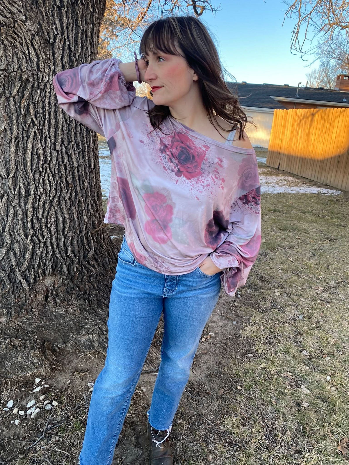 A woman wears a pink and purple floral top with blue jeans, leaning against a tree.