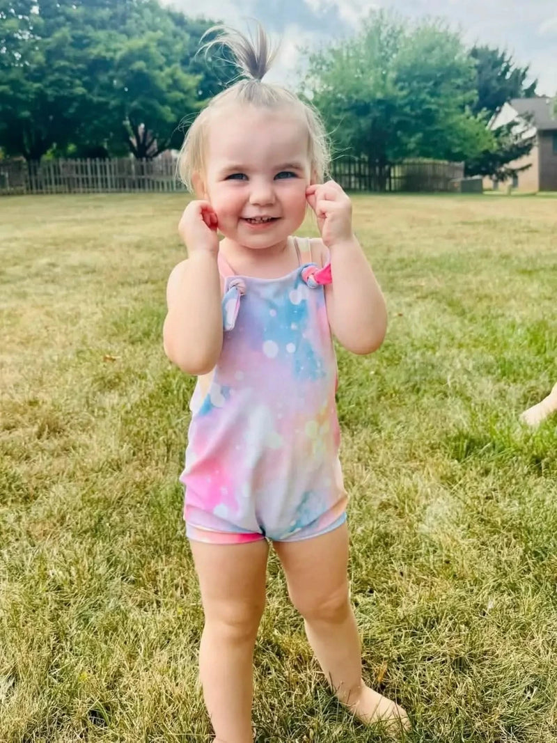 Blonde toddler girl smiling in a pink and purple sparkle print cotton romper on grass.