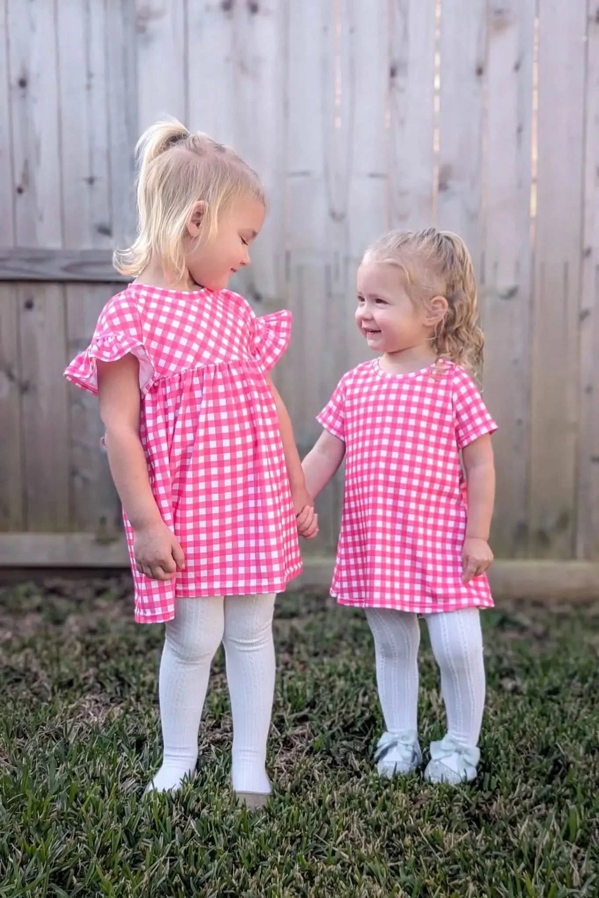 Two young girls in pink gingham dresses holding hands outdoors.