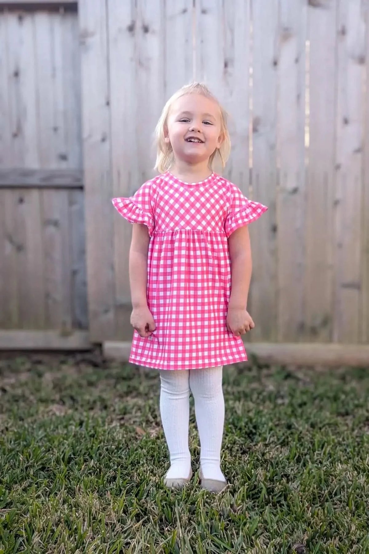 A young girl smiles while wearing a hot pink gingham dress and white tights.