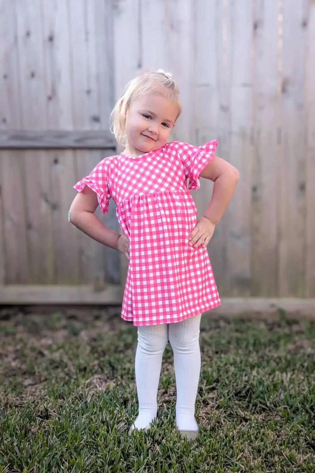 A young girl wears a hot pink and white gingham dress with white leggings.