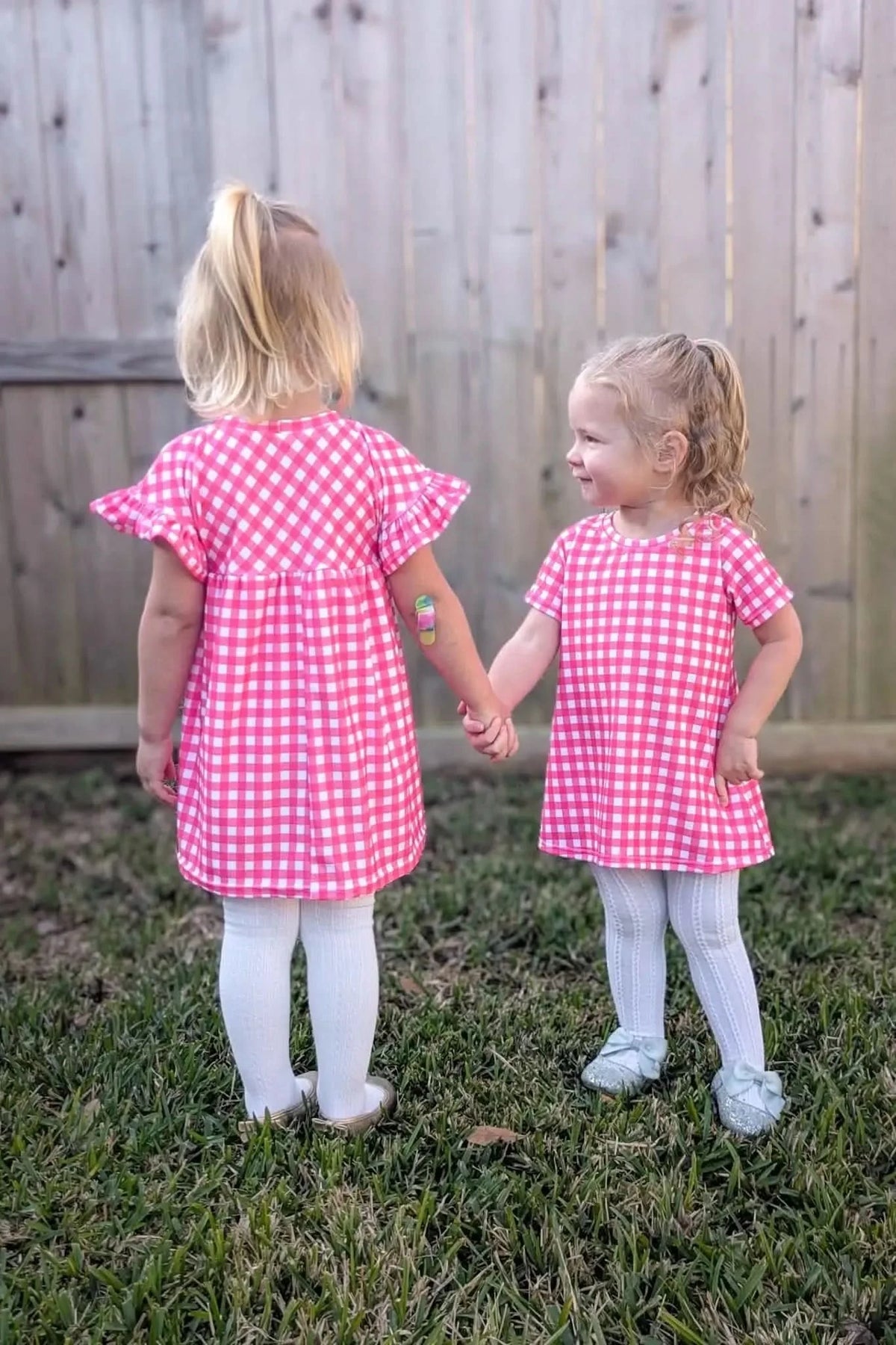 Two young girls in hot pink gingham dresses hold hands in a backyard.