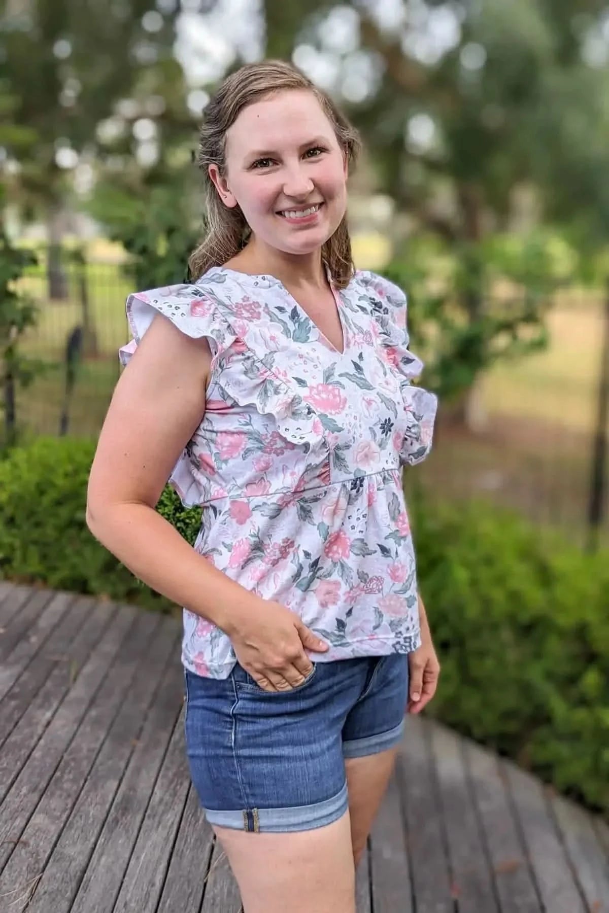 A woman wears a floral top with pink and green flowers and blue denim shorts.