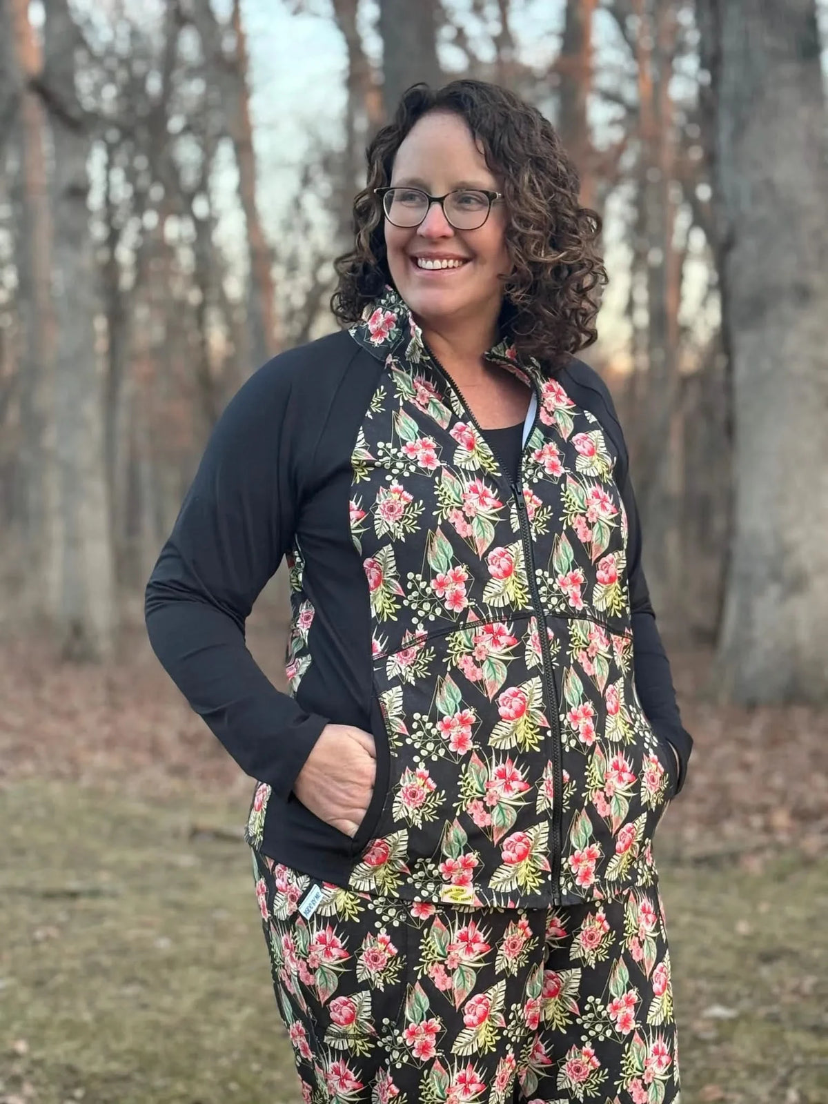 Woman modeling black zip-up hoodie with pink floral tropical print in wooded setting.