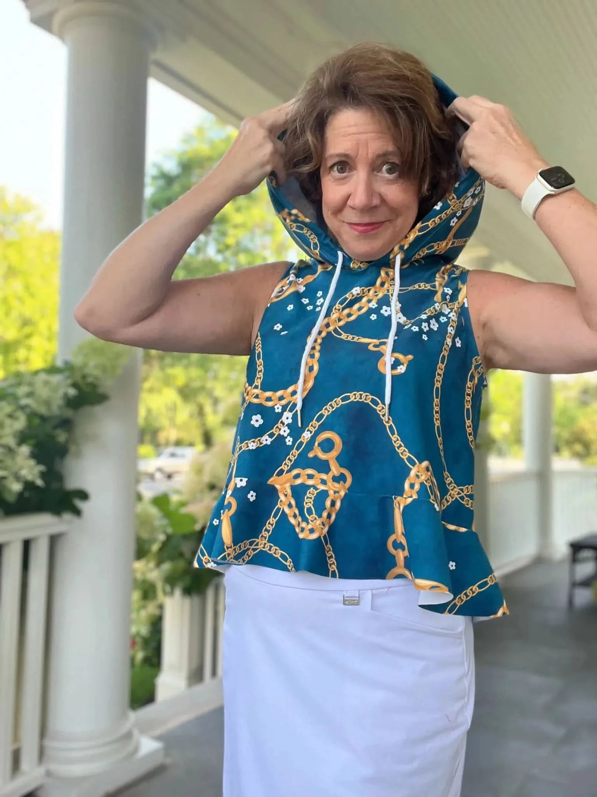 Woman modeling a sleeveless teal floral chain print hoodie top with white skirt on a porch.