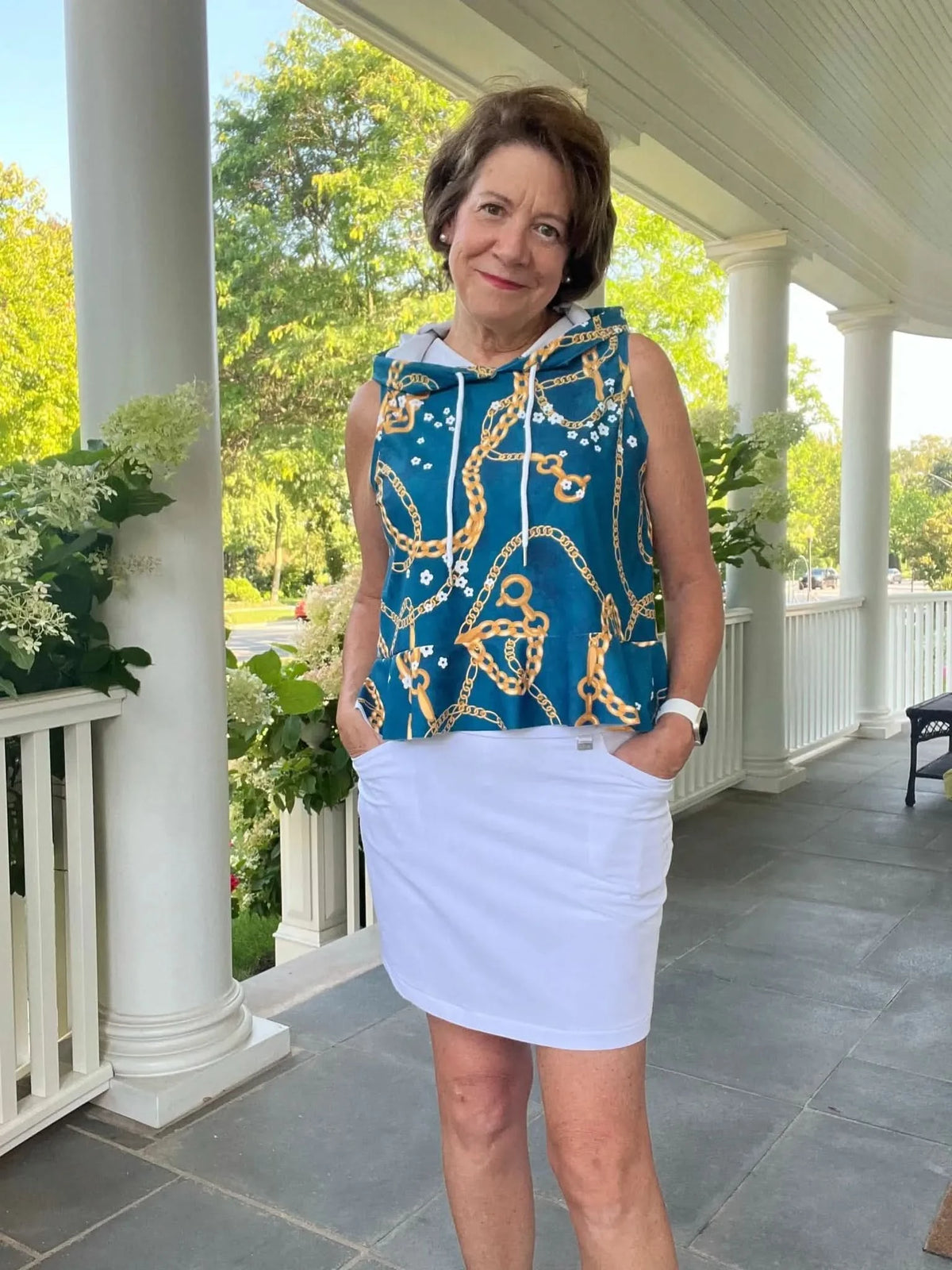 Woman modeling a sleeveless teal hoodie and white skirt made from floral chain print fabric on a porch.