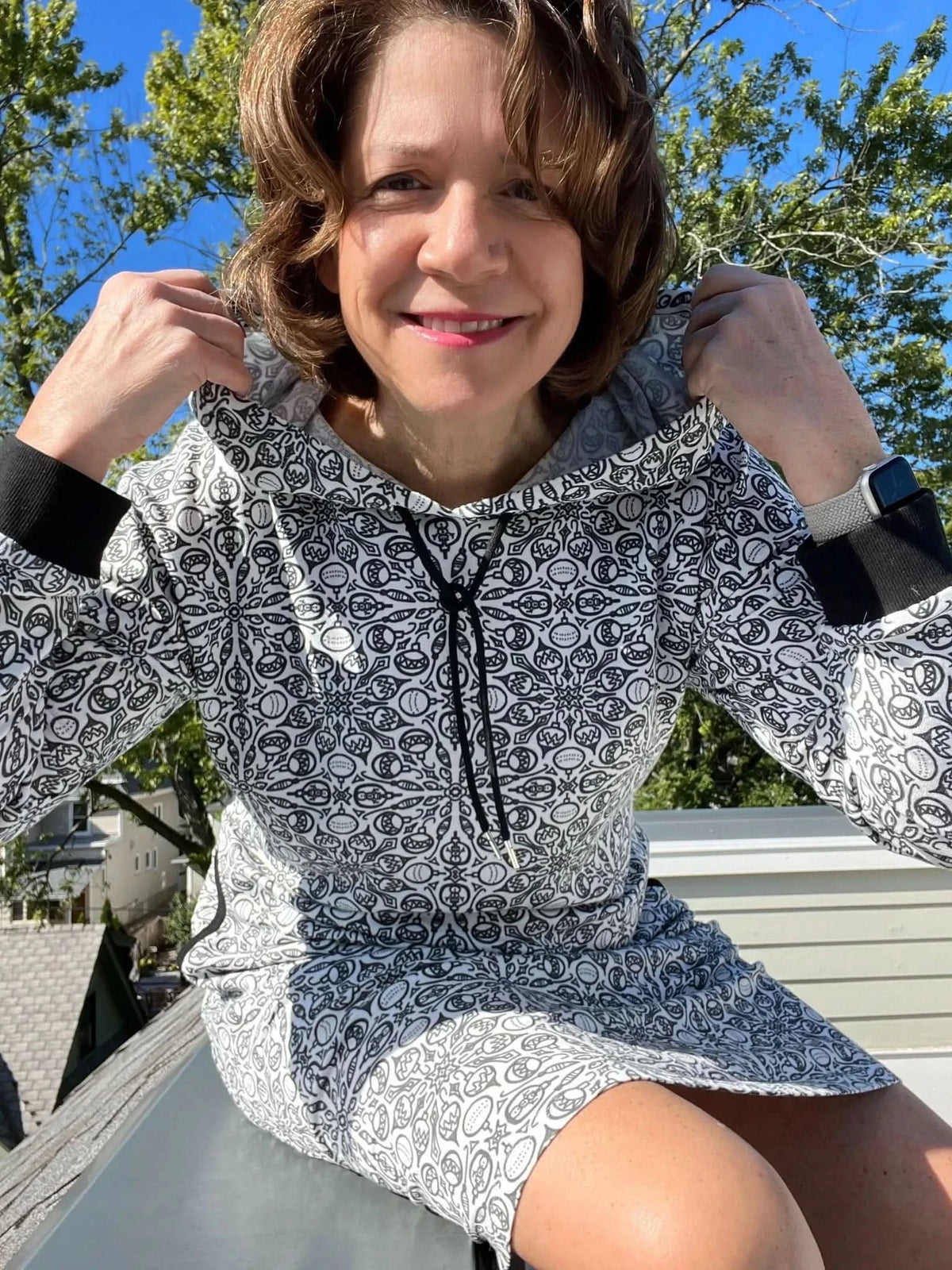 Woman modeling a white hoodie dress with black drawstrings and intricate gray print on a sunny day.