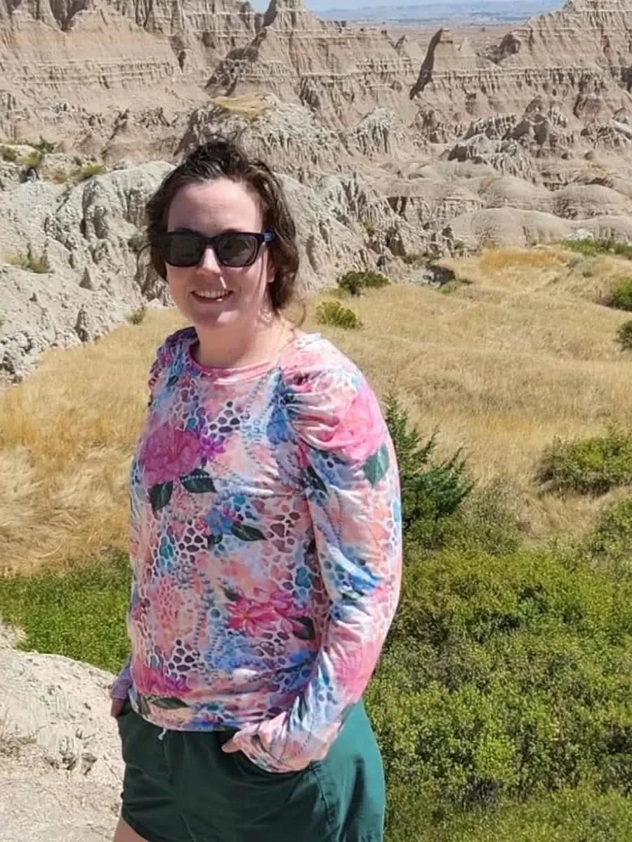Woman wearing a long-sleeved pink floral hibiscus print top with green shorts in a rocky landscape.