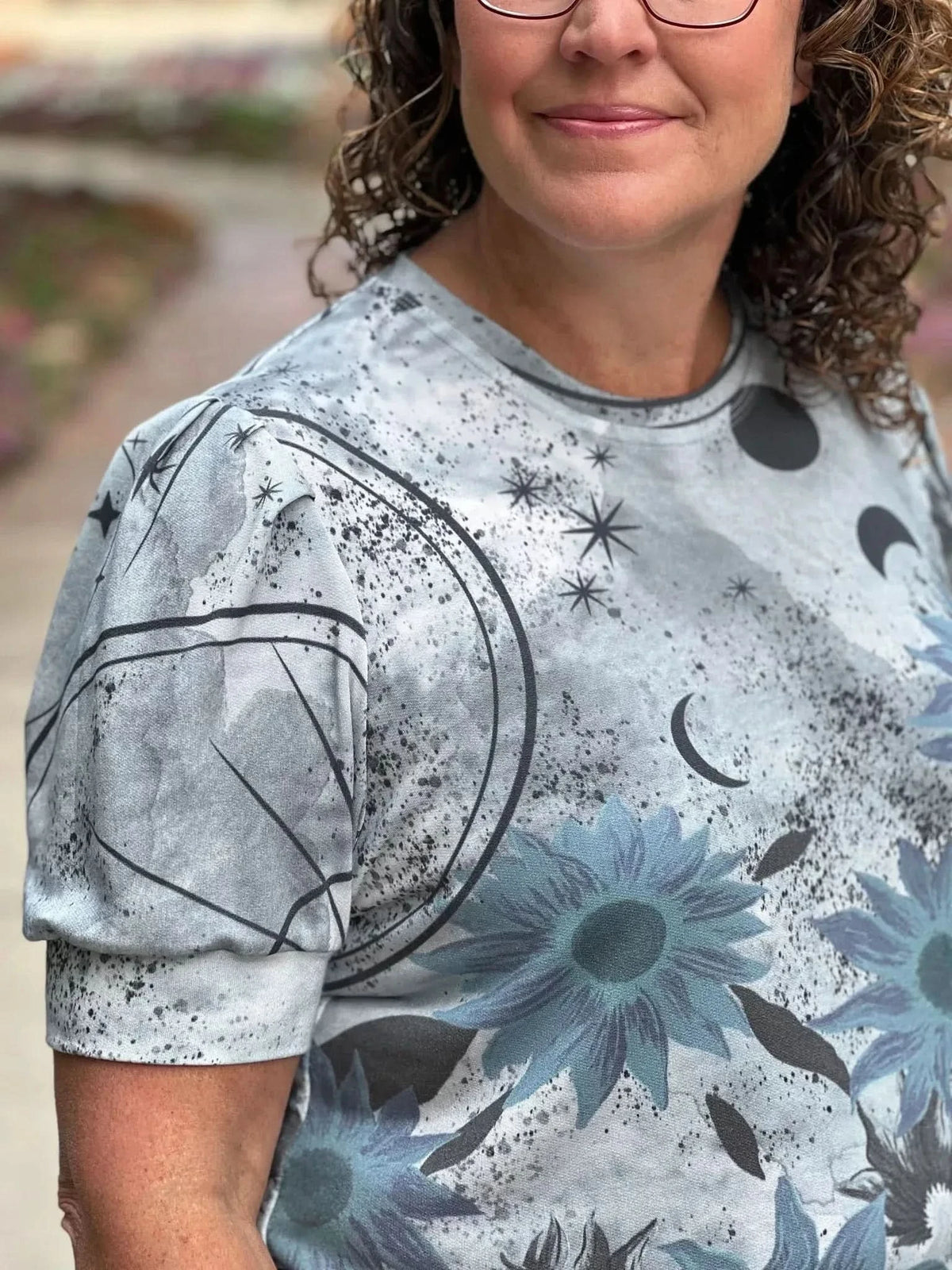 A woman wears a silver panel shirt with celestial and blue sunflower designs.
