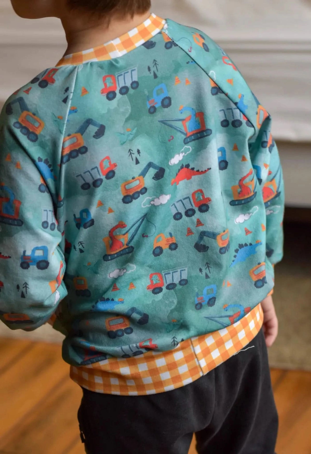 Green fabric with printed construction vehicles and dinosaurs, shown as a child's raglan sweater back.