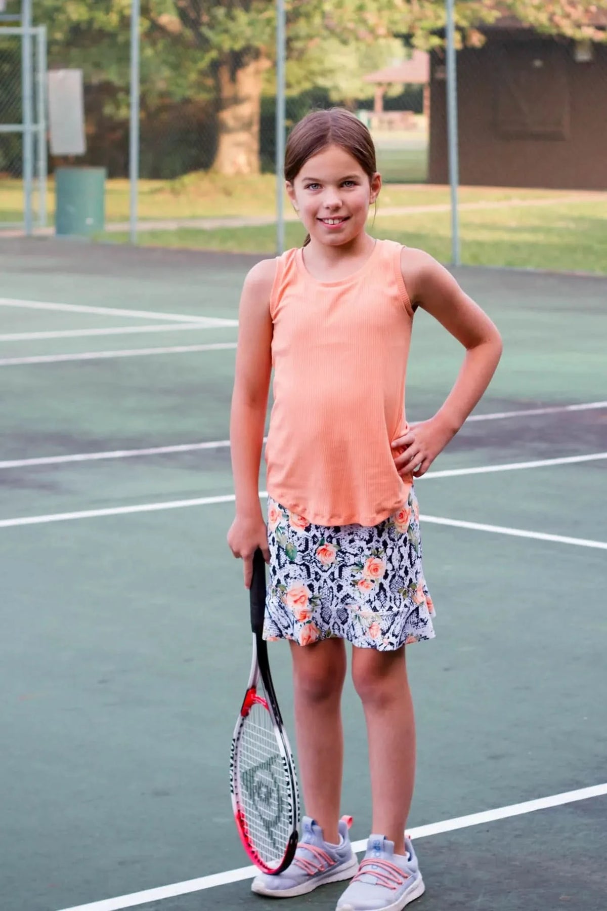 Girl in orange tank top and floral snake print skirt holding red tennis racket on court.