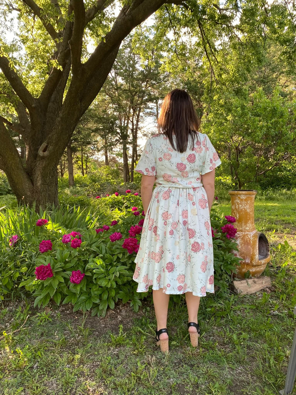 A woman in a floral dress stands with her back to the camera in a garden.