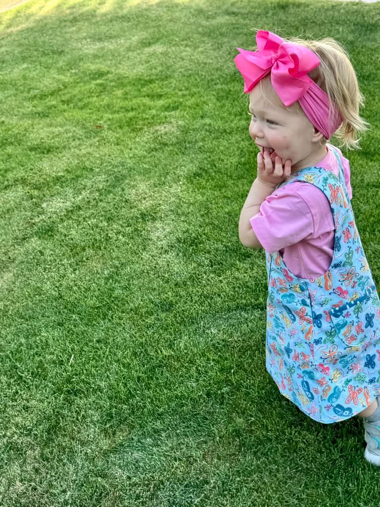 A child in a blue dress with painted butterflies stands on green grass.