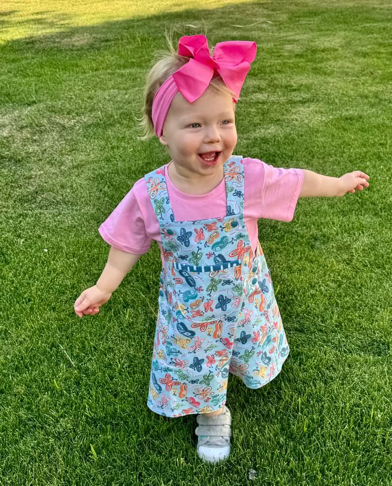 A smiling toddler wears a pink shirt and a butterfly-patterned overall dress.