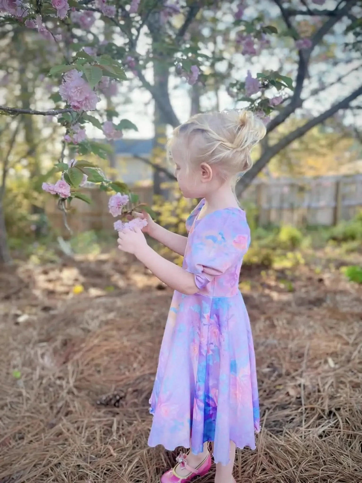 A young girl in a purple floral dress touches pink flowers on a tree branch.