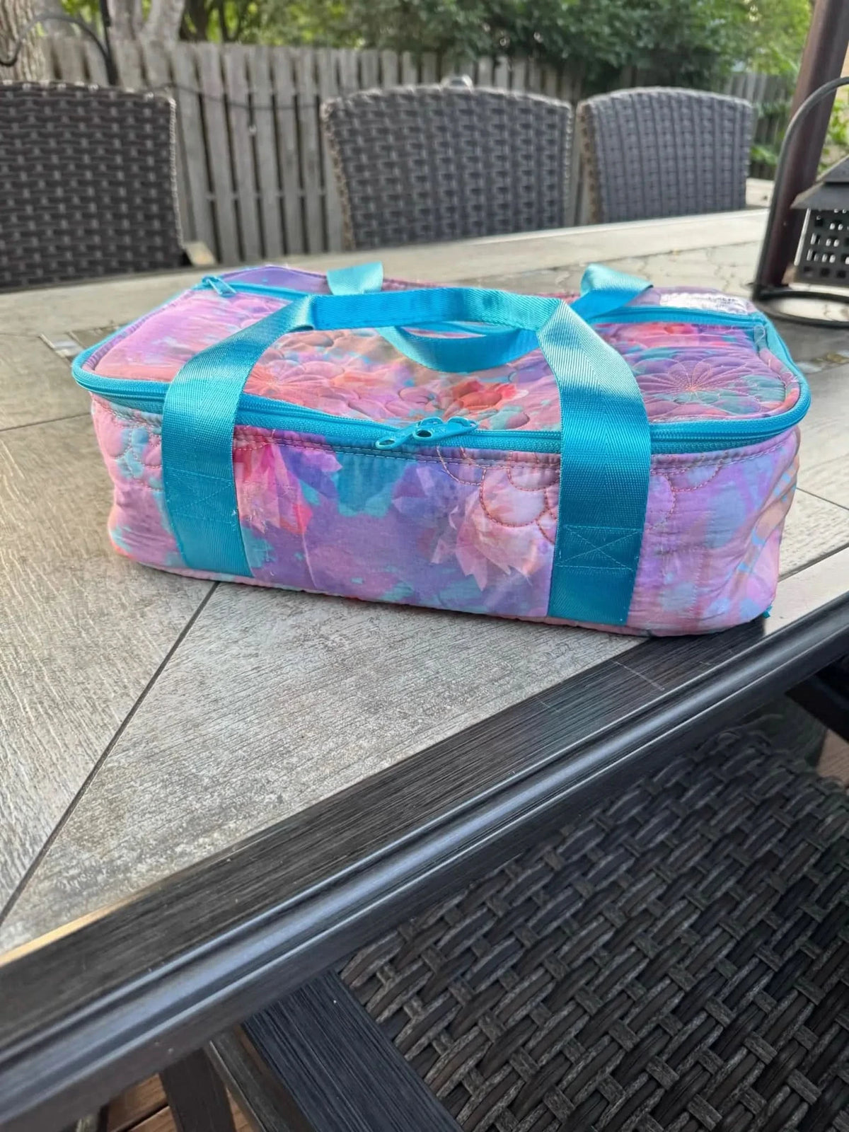 A quilted fabric bag with a floral pattern in purple, pink, and blue, with blue handles.