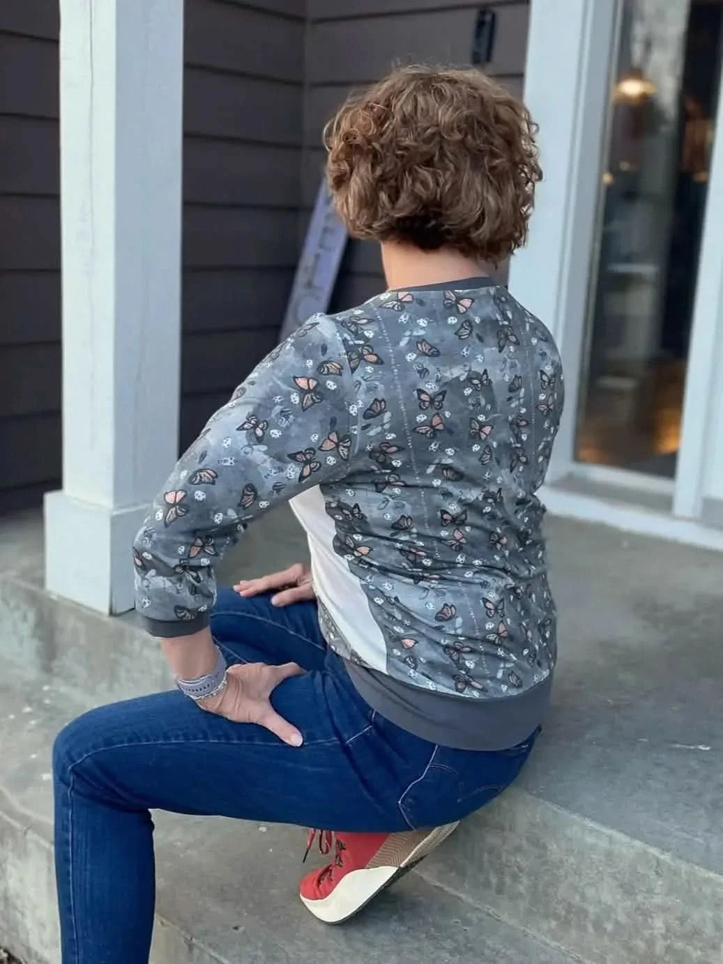 Rear view of woman wearing gray monarch butterfly print jacket and jeans, seated on porch steps.