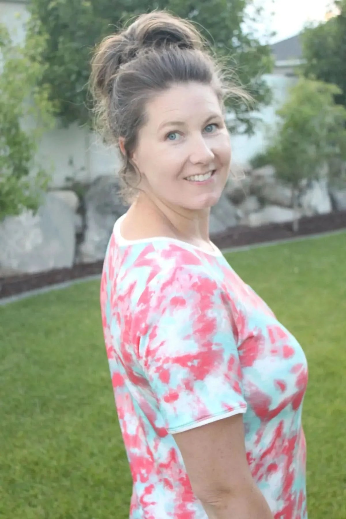 Side view of a woman modeling a pink and aqua tie-dye short-sleeve top in a backyard.