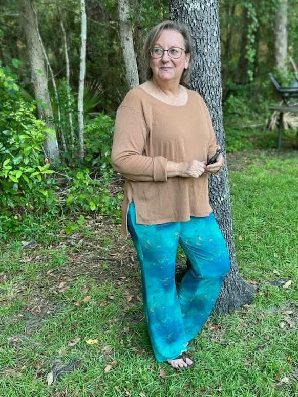 A woman in a brown top and dark teal pants with a subtle pattern stands by a tree.