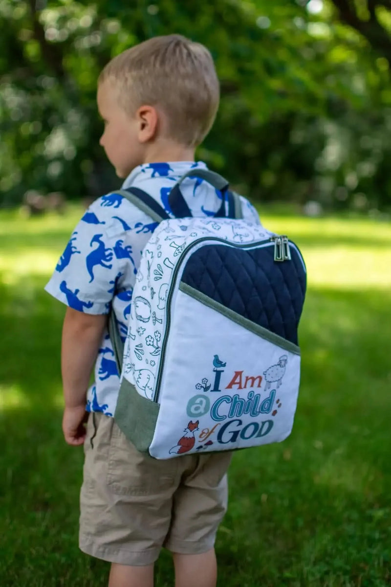 A child wearing a backpack with "I Am Child of God" text and woodland creature patterns in a park.