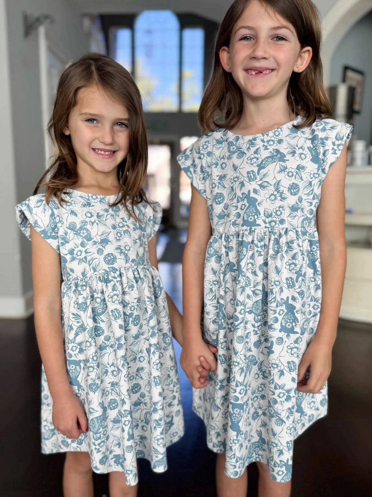 Two girls wearing matching blue Bunny Bramble print dresses in a home.