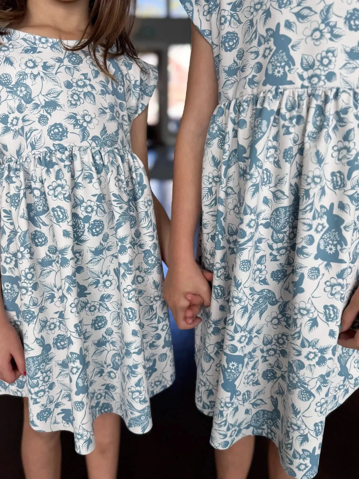 Two young girls holding hands wear matching Carolina blue dresses with bunny and bramble print.