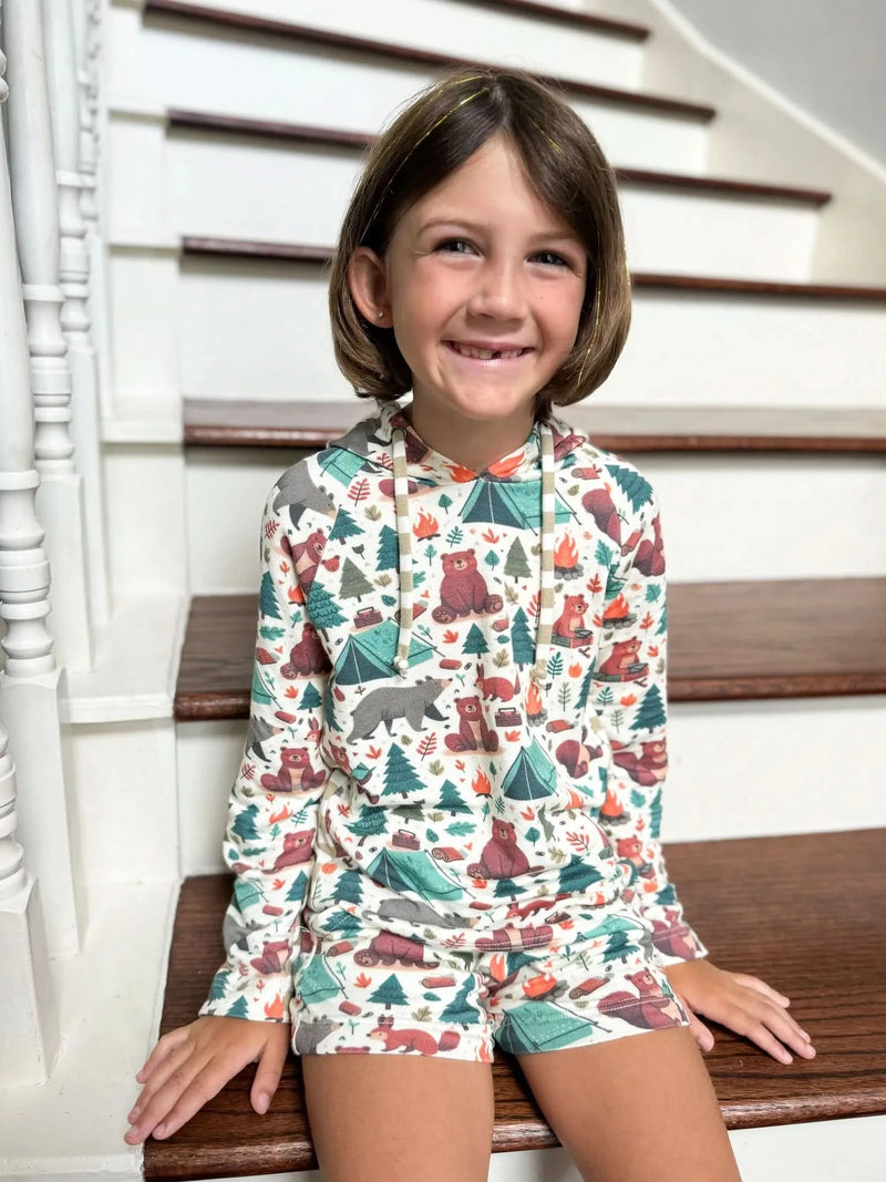 Girl wearing long-sleeve hoodie and shorts with camping bears print on wooden stairs.