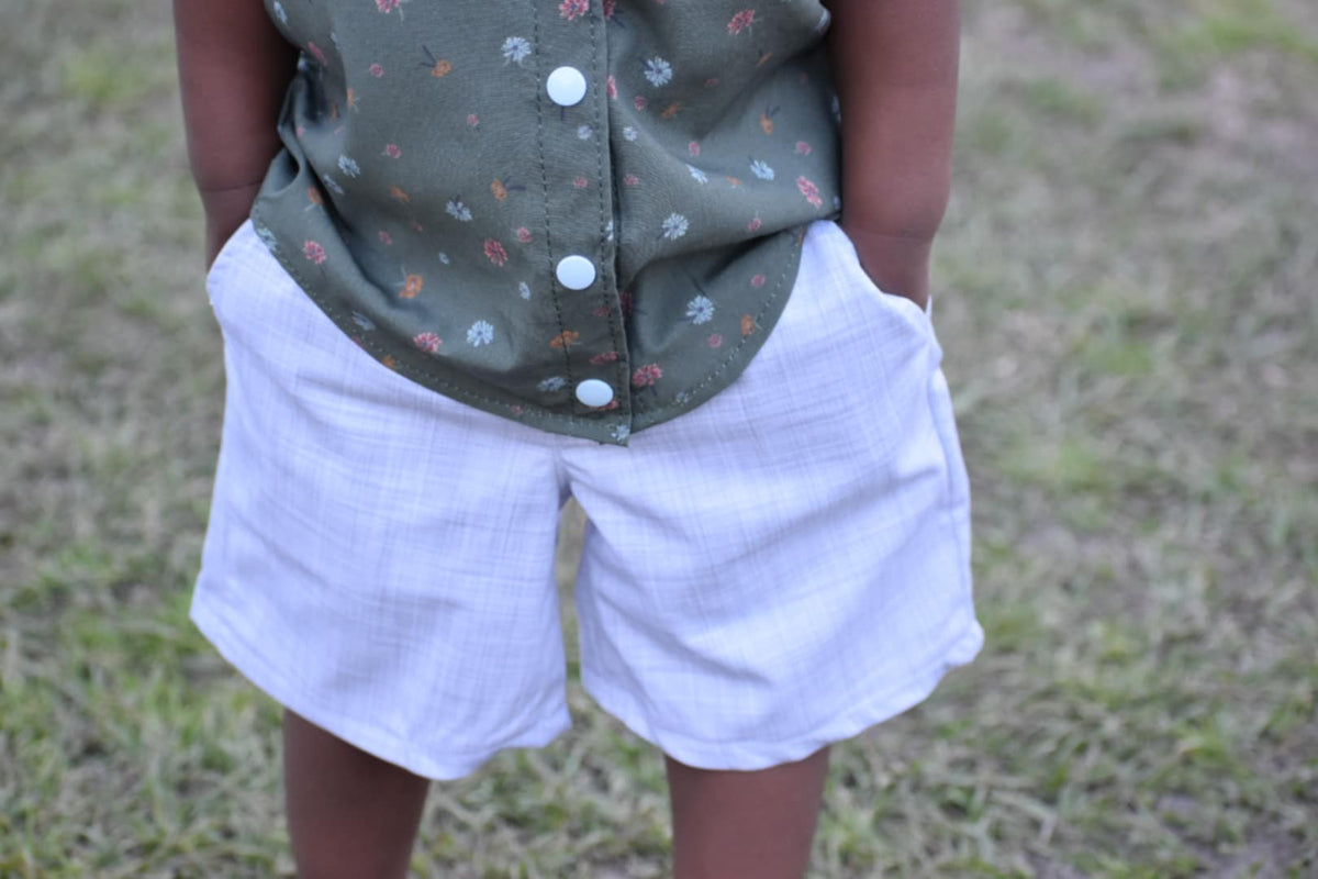 A child wears a green floral top and white plaid shorts with hands in pockets.