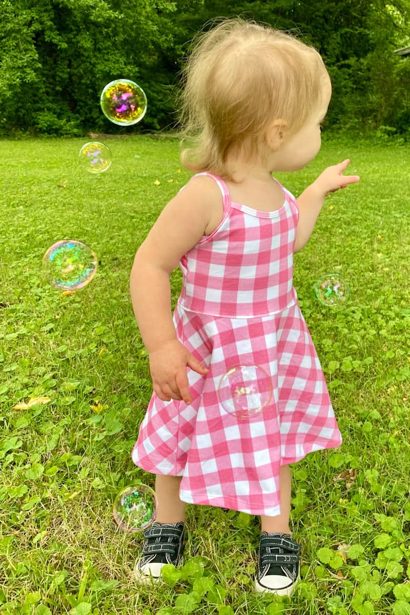 A toddler in a pink and white gingham dress plays with bubbles outdoors.