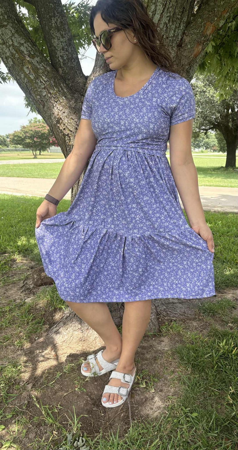 A woman in a purple floral dress and white sandals stands by a tree.