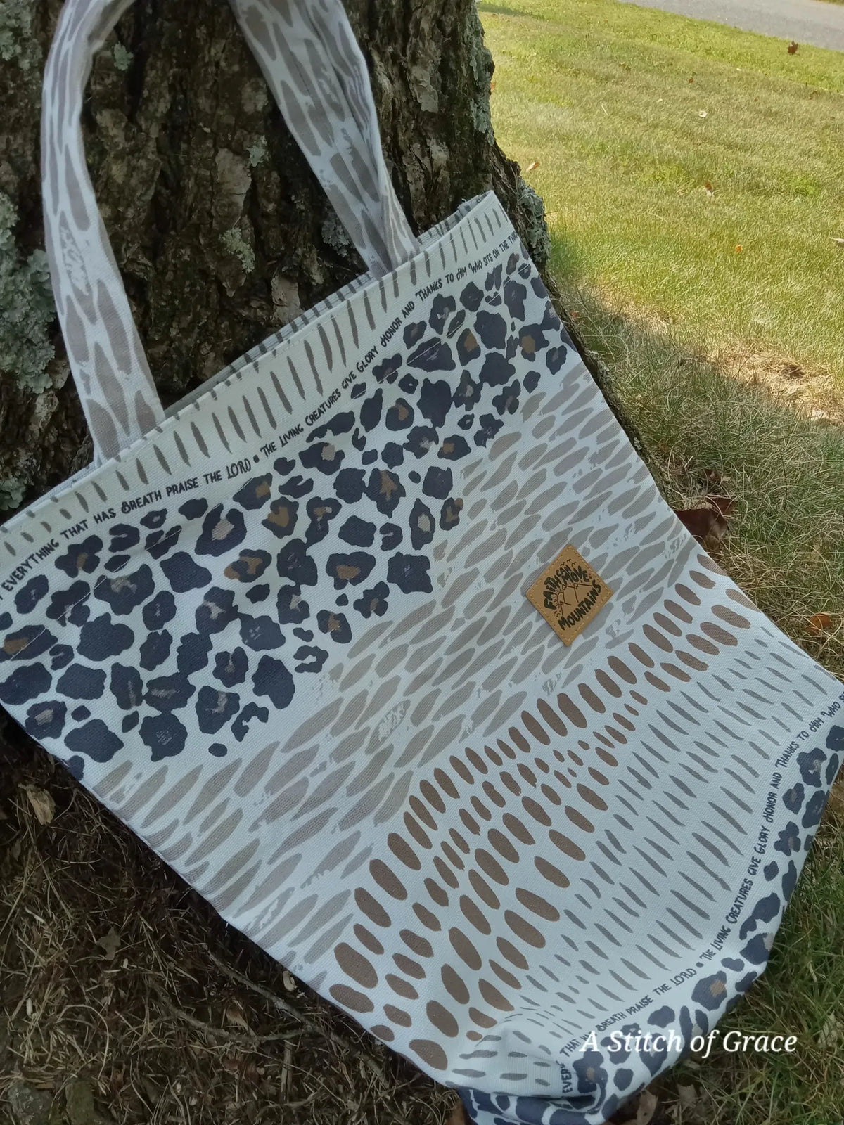 A tote bag with leopard print and abstract patterns leans against a tree.