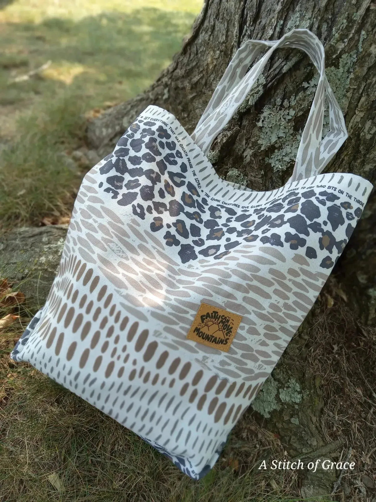 Beige tote bag with leopard print panels and "A Stitch of Grace" label, leaning against a tree outdoors.