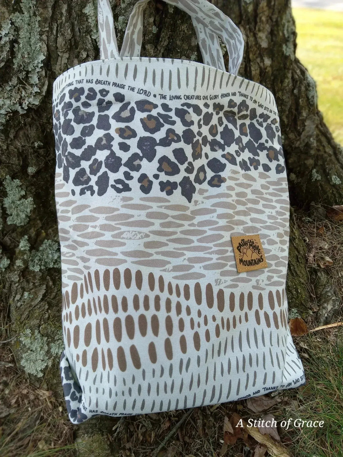 A tote bag with a faith-based print featuring animal patterns in neutral tones, hanging from a tree.