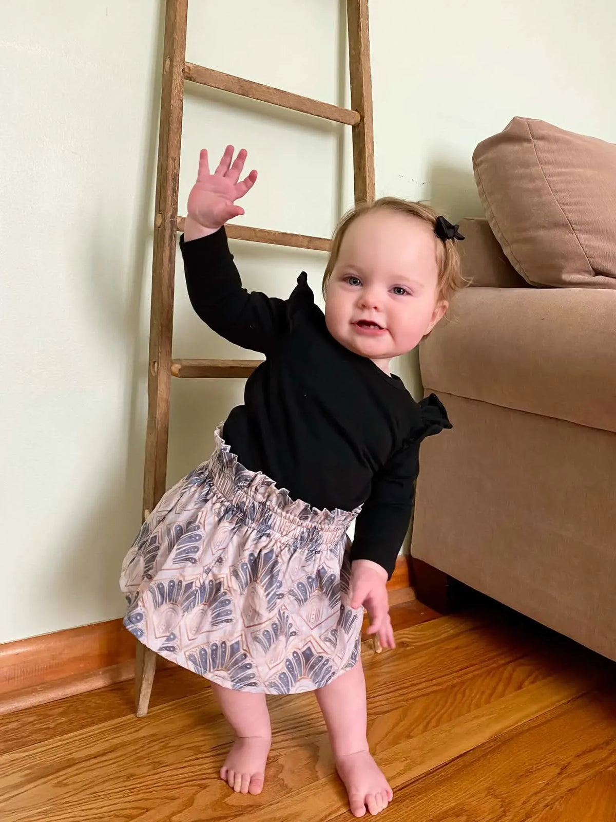 A baby in a black top and patterned skirt stands next to a wooden ladder.