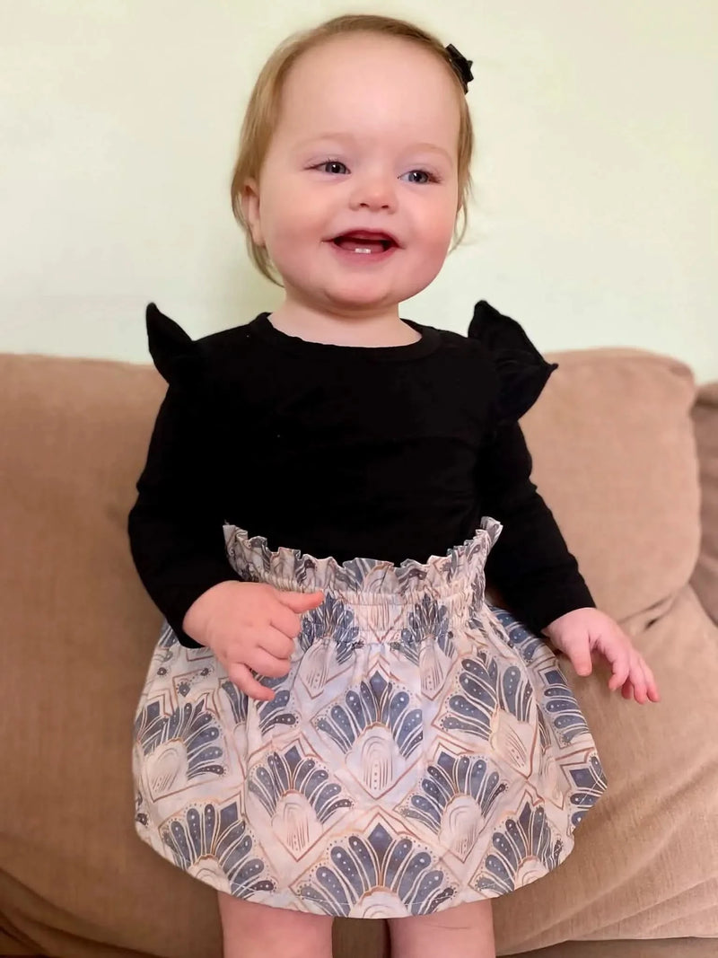 A baby in a black top and a patterned skirt with blue and gold art deco designs.