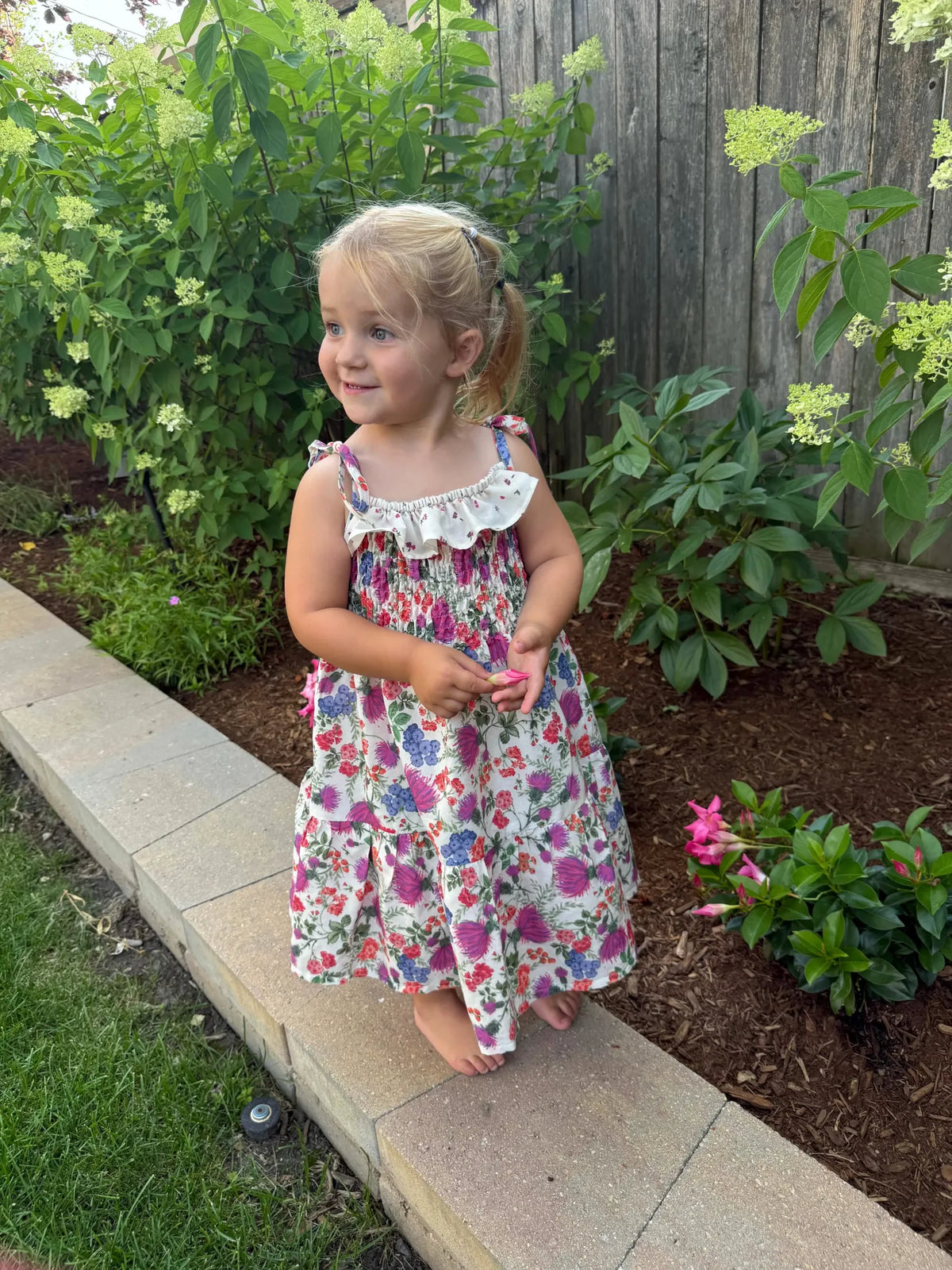 A young girl in a floral dress stands barefoot on a stone ledge.