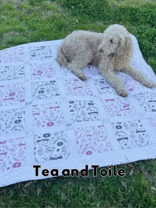 A fluffy dog lies on a white quilt with black and pink tea-themed patterns.