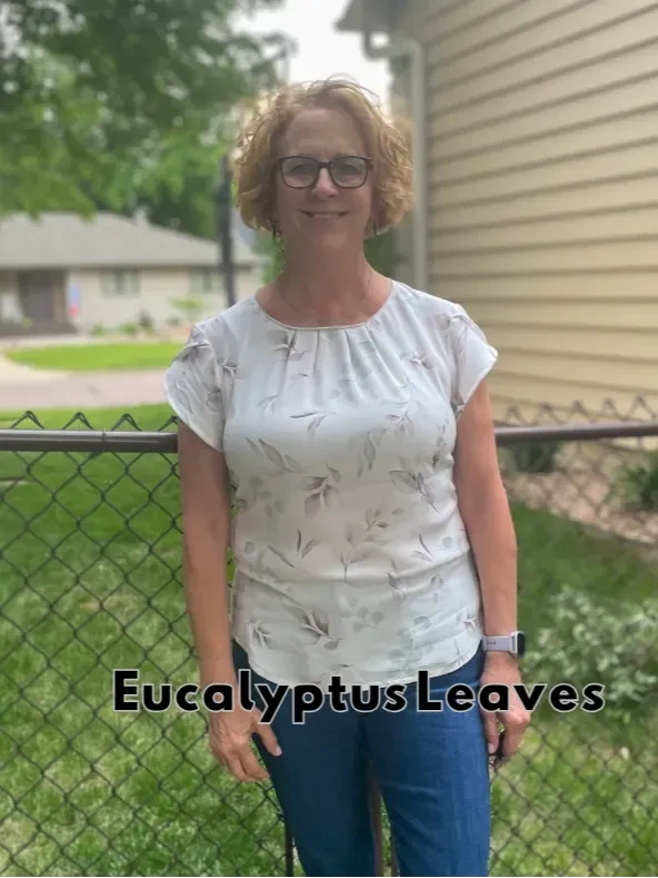 A woman in a white top with a eucalyptus leaf pattern stands in a backyard.