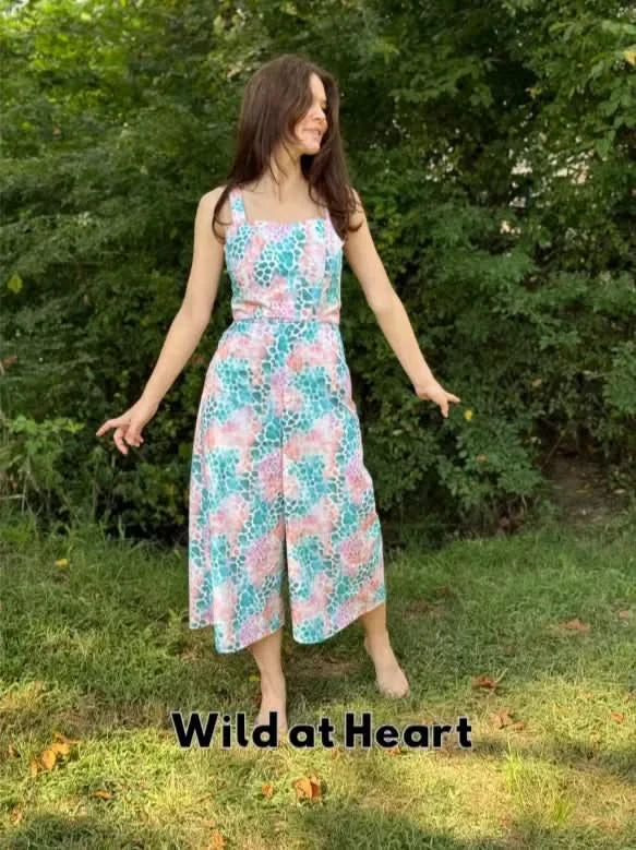 A woman in a colorful patterned jumpsuit stands barefoot in a grassy outdoor setting.