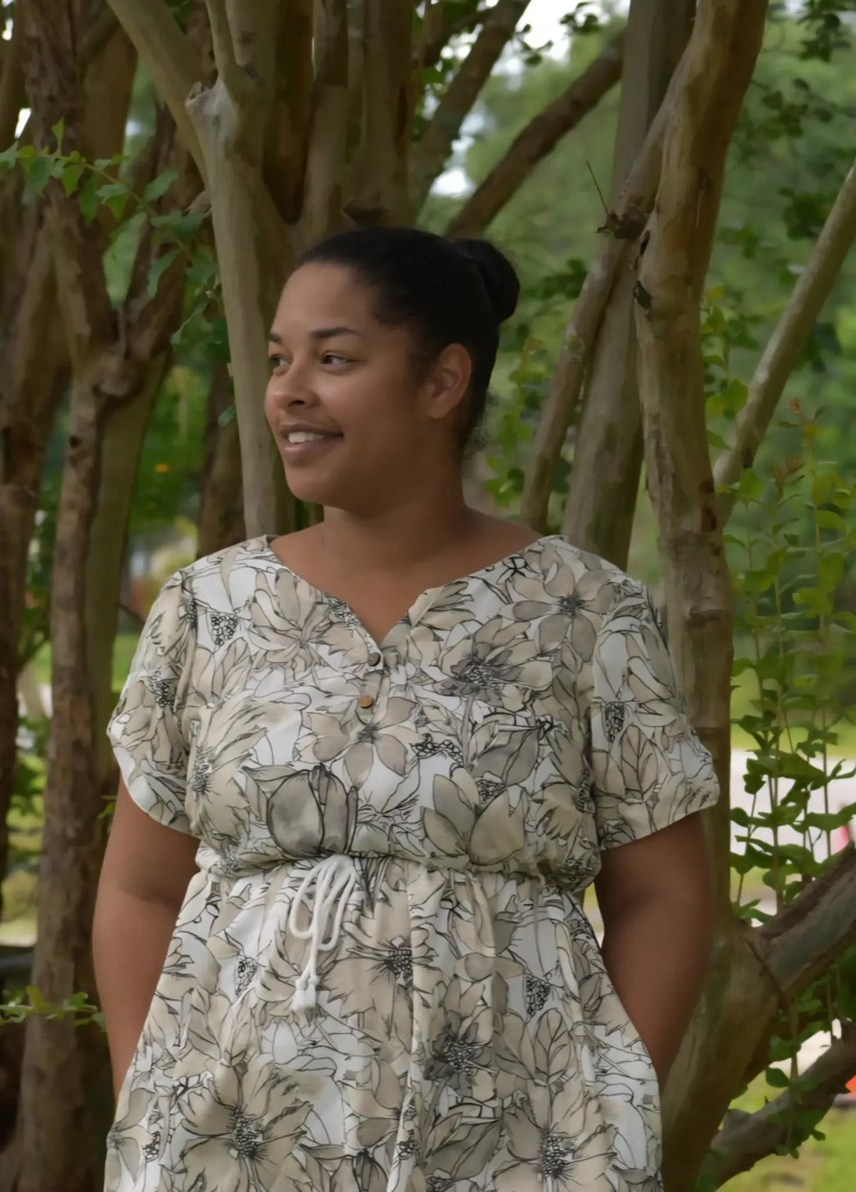 A woman in a sepia sketch floral dress stands smiling in front of trees.