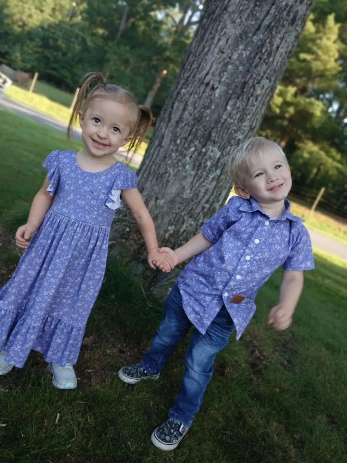 Two children in matching purple floral outfits hold hands next to a tree.