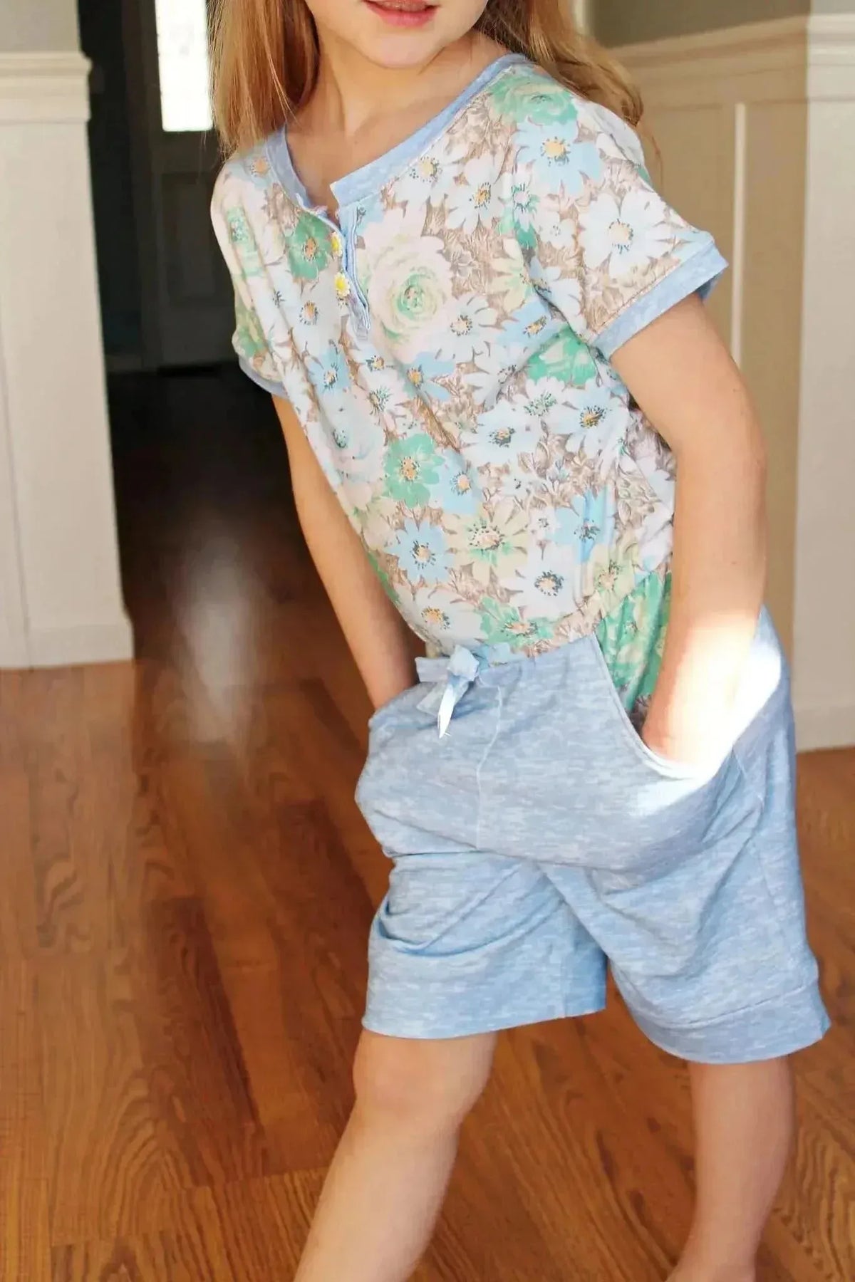 Child in a floral print top and light blue shorts with hands in pockets.
