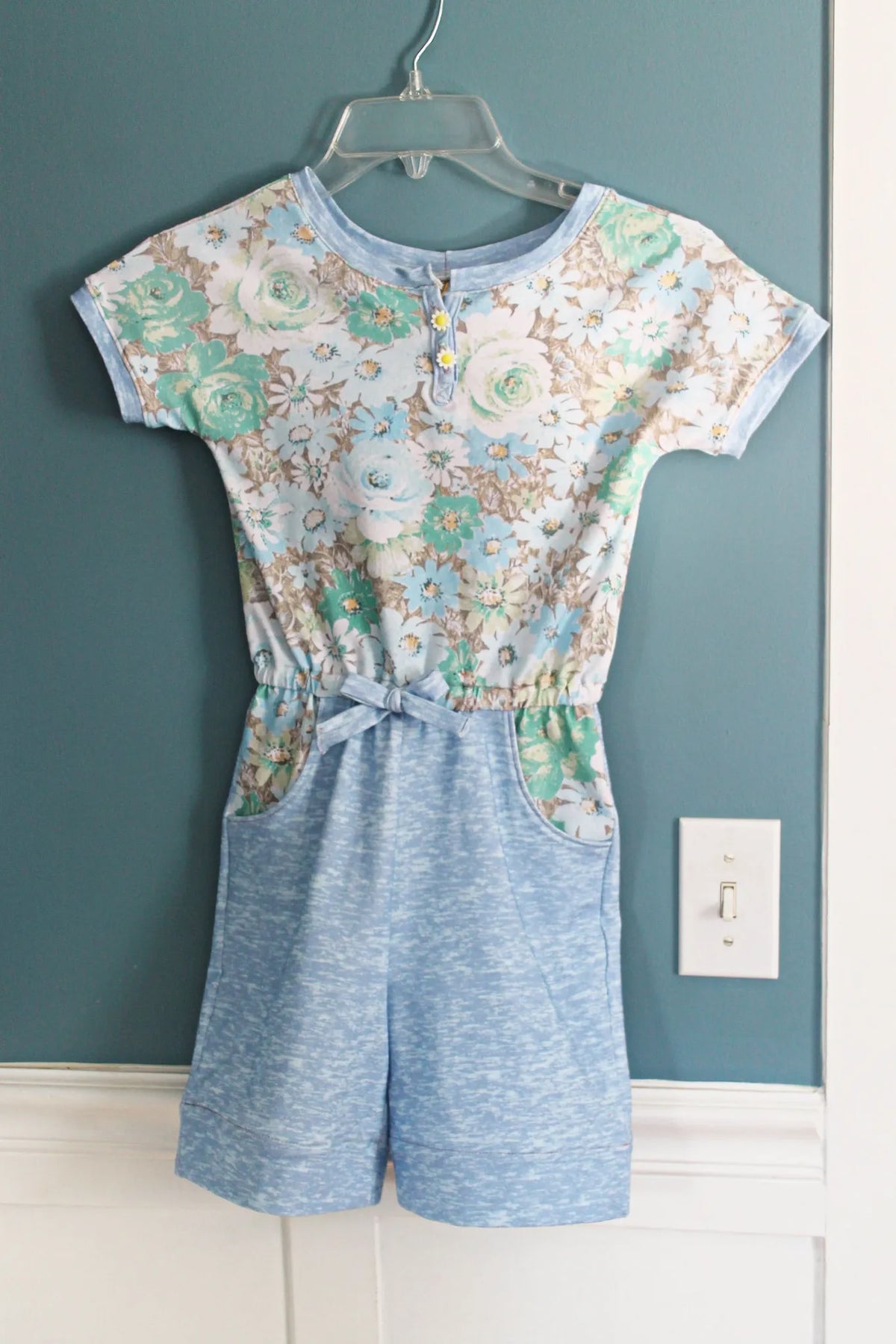 A floral print romper with a light blue top and solid blue shorts hangs on a hanger.