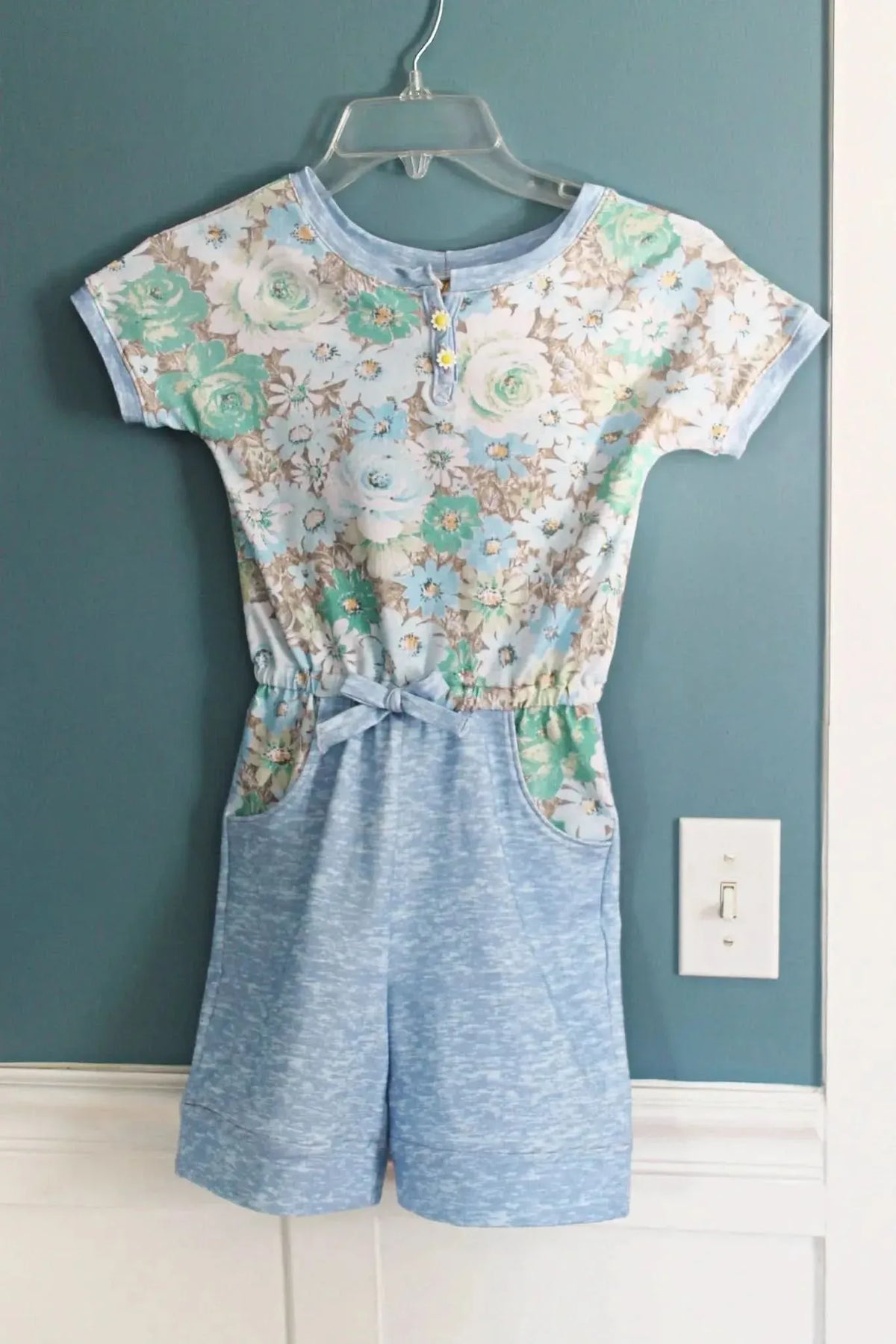 A floral print romper with a light blue top and solid blue shorts hangs on a hanger.